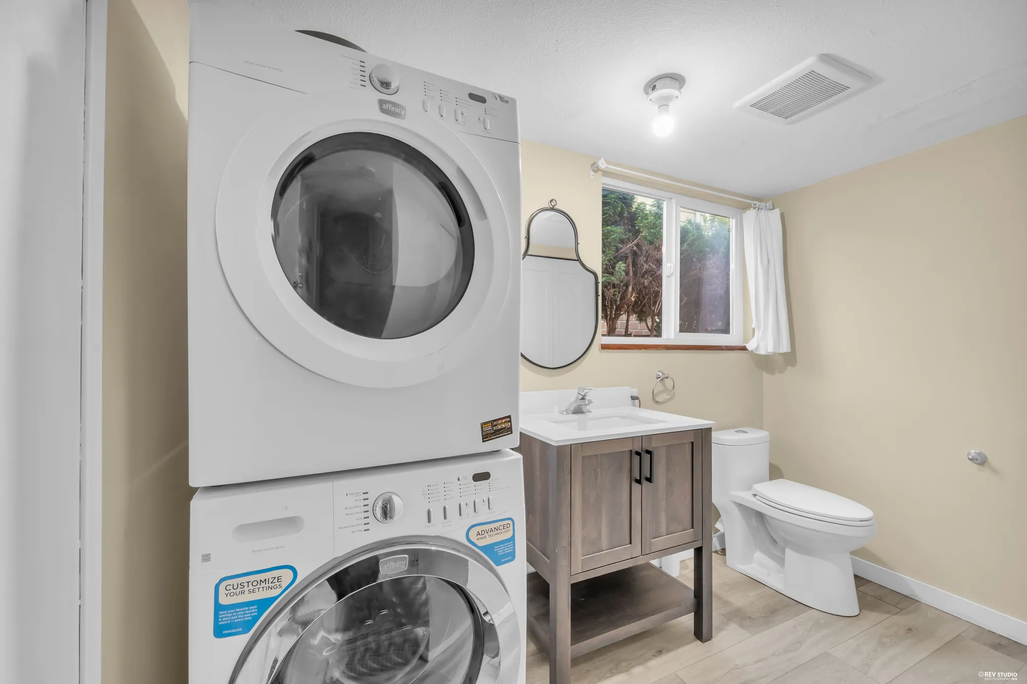 The bathroom at the side with a stacked washer and dryer, a grey vanity, a toilet and a window