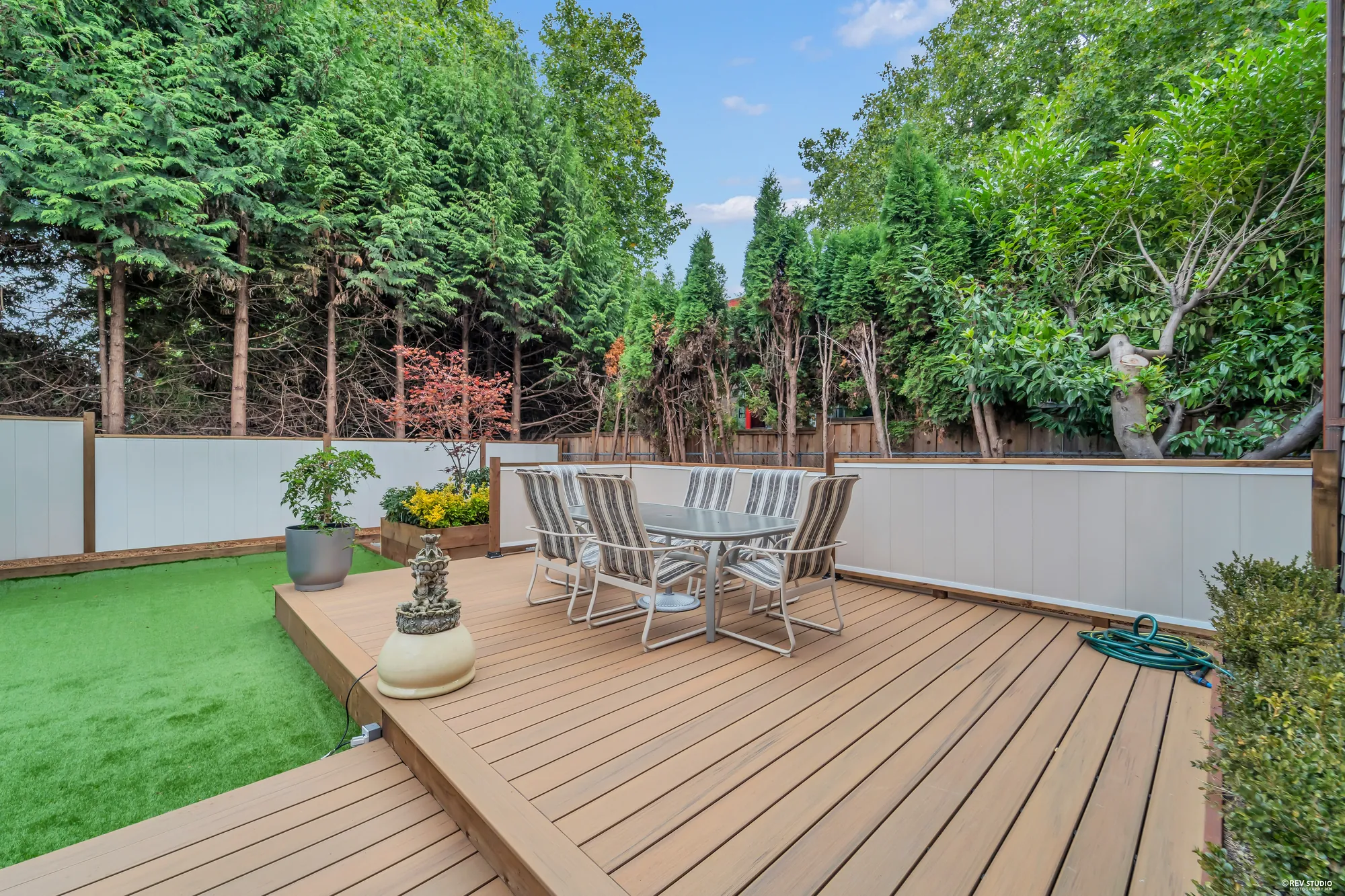 The composite deck with a table and chairs, the turf beyond and tall trees over the fence