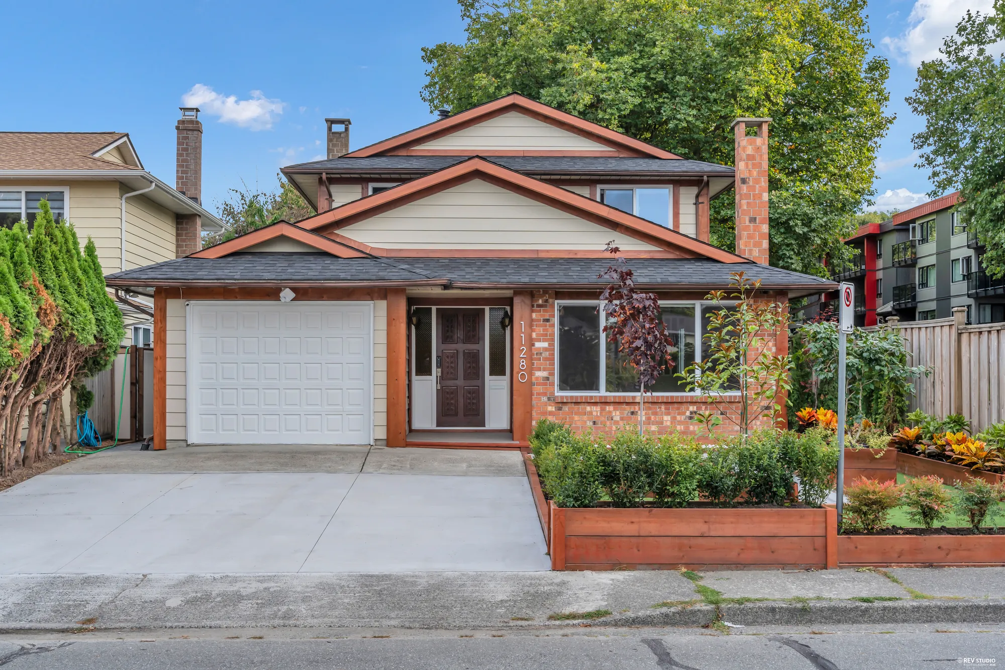 The front of the house in daylight: brick lower storey, cedar-trimmed gables, a single garage door and the raised planters along the drive