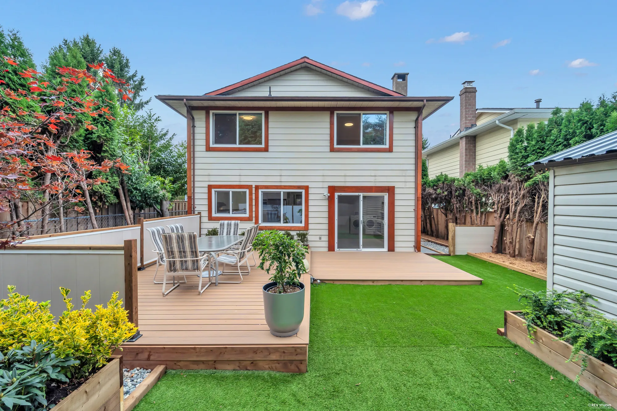 The back of the house in daylight: two storeys of cream siding with orange trim, the composite deck and dining set below, turf and a shed to the right